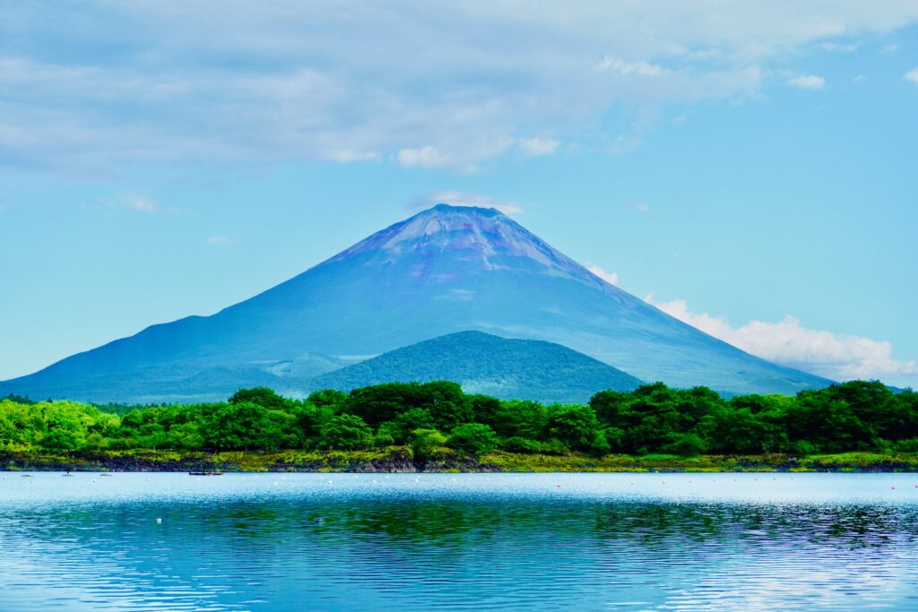 Mt. Fuji without snow on a clear summer day, rising above a calm lake and deep green forest