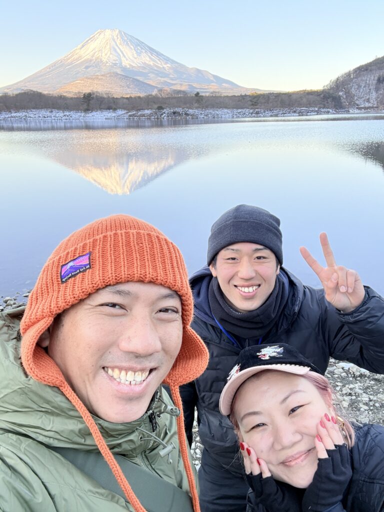 A guide and guests taking a selfie beside a calm lake reflecting snow-capped Mt. Fuji