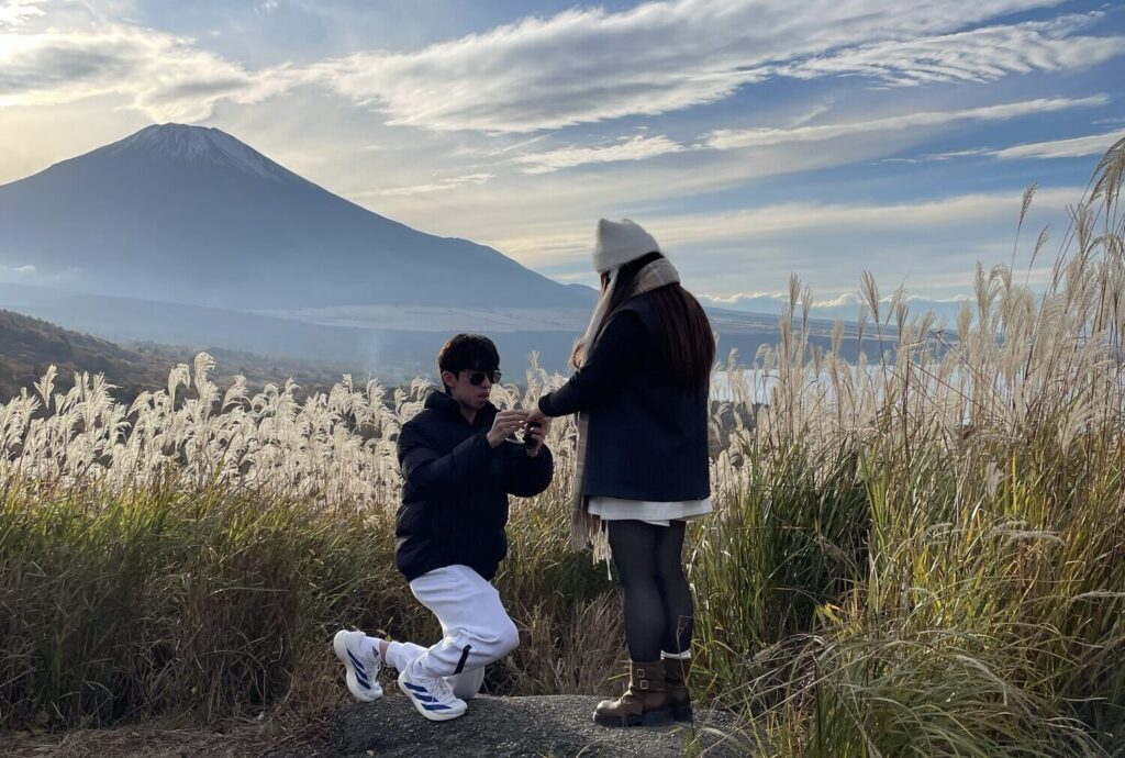 A man kneeling to propose among pampas grass at a hidden viewpoint facing Mt. Fuji